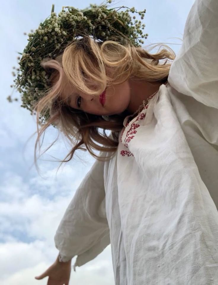 Une femme avec une couronne de fleurs se tient debout, elle ne porte pas de maquillage mais sa peau est éclatante et sans défaut, ses cheveux sont blonds et traduisent sa beauté naturelle et sa bonne santé. Elle ouvre les bras, comme pour dire oui au monde, oui à la nature, et oui à l'expansion de sa conscience.